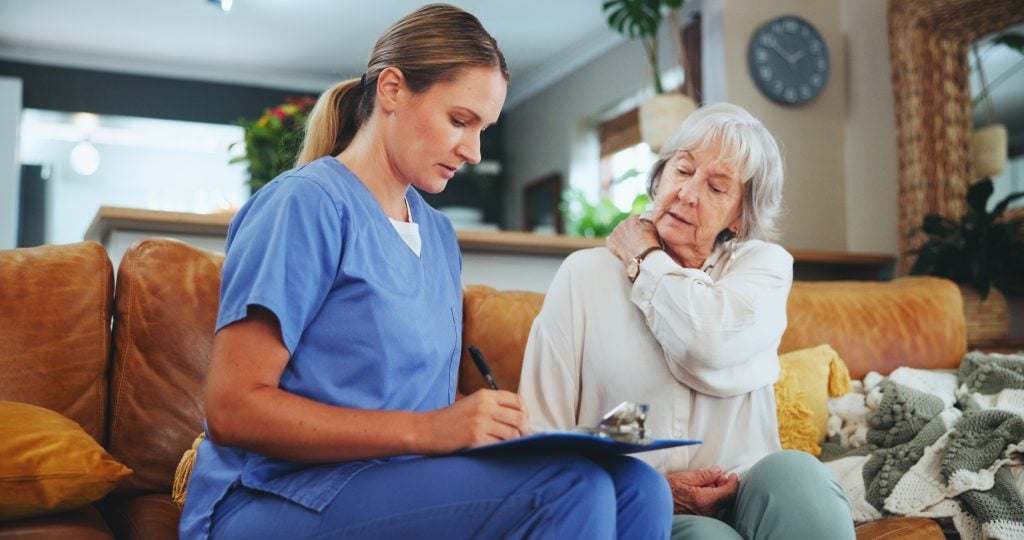 Female caregiver in blue scrubs writing on a clipboard while an elderly woman looks on in a home setting.