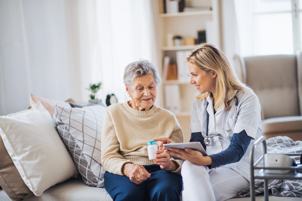 Smiling female healthcare professional with a stethoscope showing a tablet to an elderly woman holding medication in a home setting.